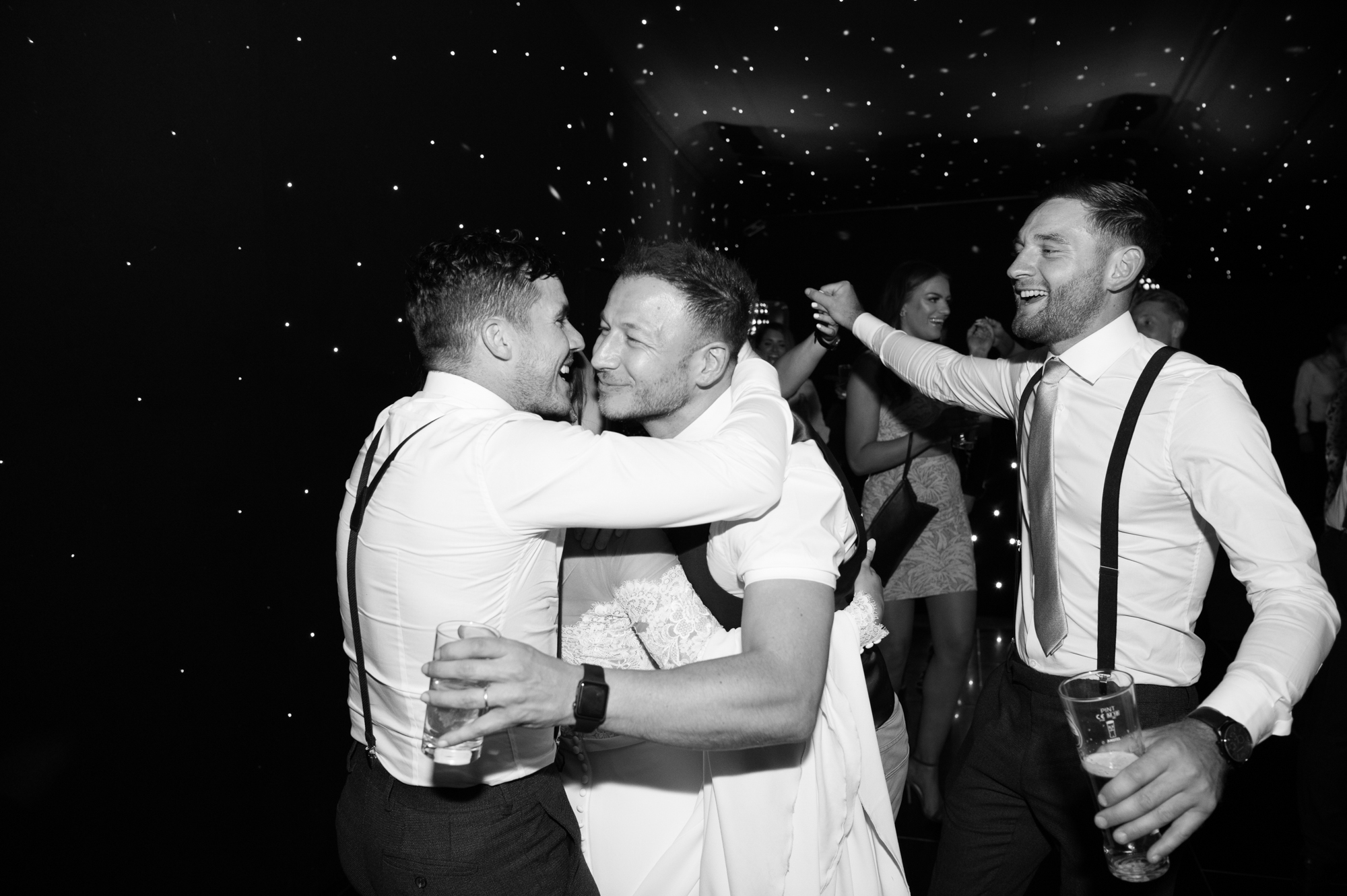 A black and white shot of male guests wearing braces dancing at a wedding